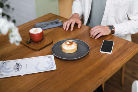 cropped view of man near lemon tart, cappuccino, menu and smartphone with blank screen on table