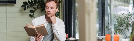 young curly man in wireless earphones reading book in cafe, banner