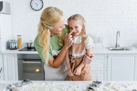 woman touching nose of cheerful daughter near kitchen table with dough and cookie cuttersの写真素材