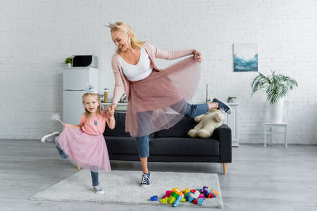 joyful mom and daughter in toy crowns and tulle skirts dancing at homeの写真素材