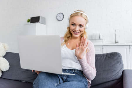cheerful woman in headphones waving hand during video chat on laptop