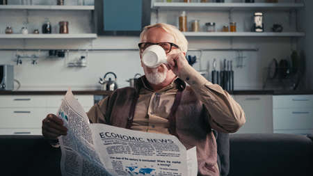 Senior man in eyeglasses holding newspaper while drinking tea at homeの写真素材