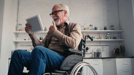 Smiling disabled pensioner in wheelchair showing thumb up during video call at homeの写真素材