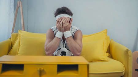 displeased football fan in sportswear covering face while watching tv on sofaの写真素材