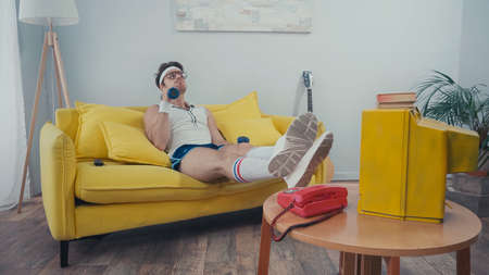 Sportsman lifting dumbbells while sitting on couch near retro tv in living roomの写真素材