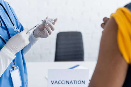 partial view of doctor in latex gloves holding vaccine near blurred african american man in...