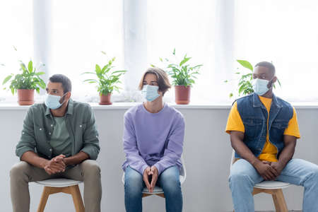 young multiethnic men in medical masks sitting in line while waiting for vaccinationの写真素材