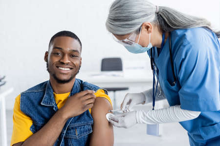asian doctor in medical mask vaccinating happy african american man in clinic