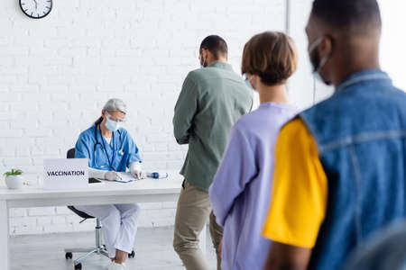 blurred multiethnic patients in medical masks standing in line near mature asian doctor writing in vaccination centerの写真素材