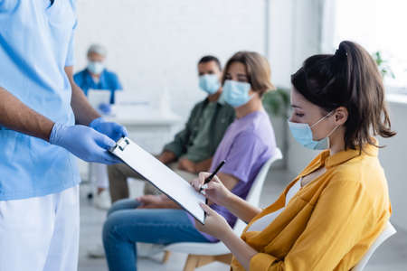 pregnant woman in medical mask signing paper on clipboard in hands of african american doctor, vaccination conceptの写真素材