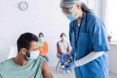 mature asian physician in medical mask holding syringe and jar with vaccine near young man and...