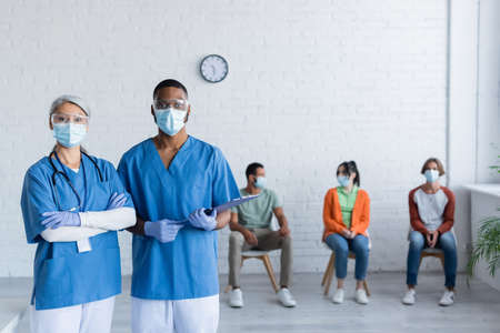 interracial doctors in medical masks and goggles looking at camera near patients on blurred backgroundの写真素材
