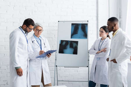 middle aged asian doctor pointing at clipboard near lungs fluorography and multiethnic colleaguesの写真素材