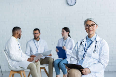 middle aged asian physician with clipboard looking at camera near multiethnic colleagues talking on blurred backgroundの写真素材