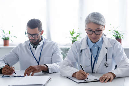 young and mature interracial doctors in eyeglasses writing on clipboardsの写真素材