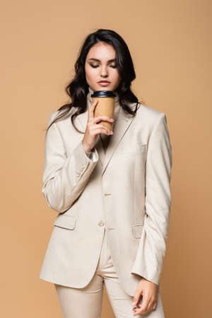 young pretty woman in turtleneck and suit holding paper cup isolated on beigeの写真素材