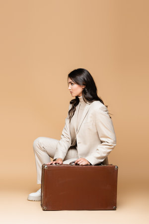 side view of young traveler in suit posing near vintage suitcase on beigeの写真素材