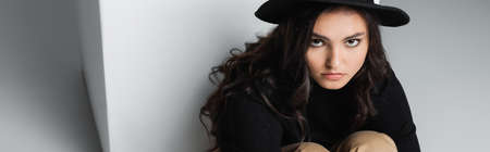young model in fedora hat looking at camera while sitting near white cube on grey, bannerの写真素材