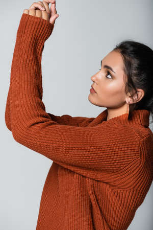 young woman in earring and knitted sweater looking up isolated on grey