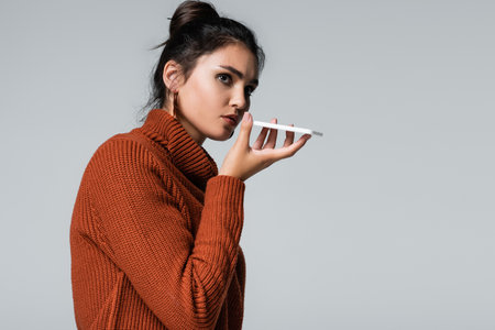 young woman in warm sweater recording voice message on smartphone isolated on greyの写真素材