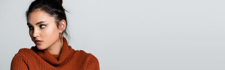 pretty young woman in earrings and knitted sweater looking away isolated on grey, banner