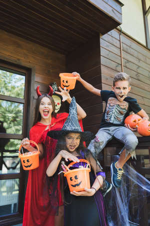 kids in creepy halloween costumes showing scary gestures while holding buckets with sweets on cottage porchの写真素材