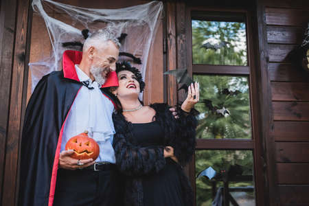 happy man in vampire costume holding carved pumpkin and embracing wife on cottage porch with halloween decorationの写真素材