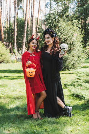 cheerful mom and daughter in demon and vampire halloween costumes posing with skull and bucket of...