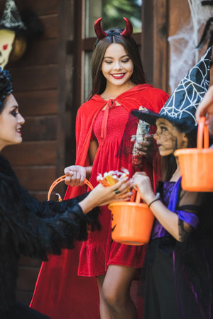 mom giving candies to happy daughters in devil and witch halloween costumesの写真素材