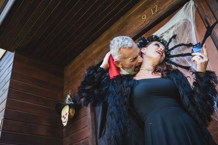 low angle view of man in halloween vampire costume biting neck of wife on cottage porchの写真素材