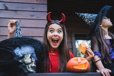 amazed girl in devil costume near sister in witch hat and carved pumpkin on cottage porchの写真素材