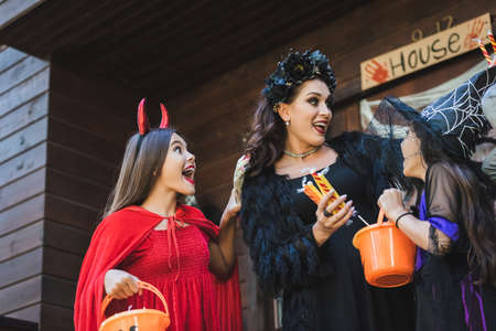 excited woman holding candies near daughters in devil and witch halloween costumesの写真素材