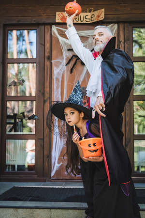 man in halloween vampire costume holding carved pumpkin in raised hand near daughter in witch hatの写真素材