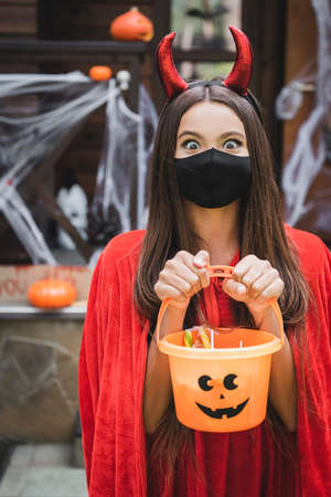 excited girl in devil halloween costume and black medical mask holding bucket with sweets while...