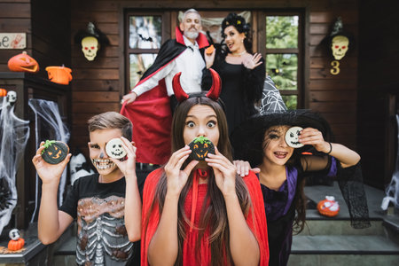 excited kids in scary costumes having fun with halloween cookies while mother waving hand on blurred...