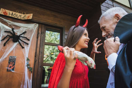 cheerful girl in devil halloween costume scaring dad with toy hand near cottage with decoration