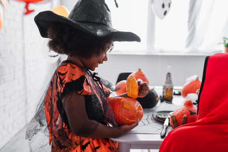 happy african american girl in pointed hat holding carved pumpkin near brotherの写真素材