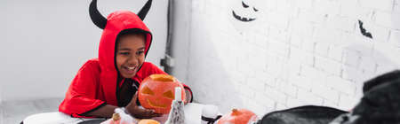 happy african american boy in halloween costume holding carved pumpkin at home, bannerの写真素材