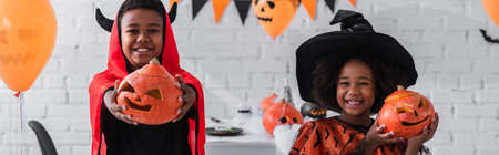 cheerful african american kids in halloween costumes holding carved pumpkins at home, bannerの写真素材