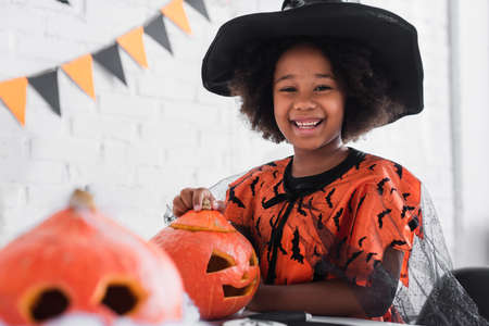 happy african american girl in witch halloween costume near caved pumpkinsの写真素材