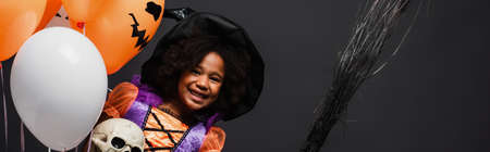 happy african american girl in halloween costume holding broom and balloons isolated on black, bannerの写真素材