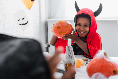 happy african american boy in halloween costume holding carved pumpkin near blurred sisterの写真素材
