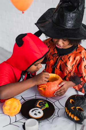 african american kids in halloween costumes carving pumpkin near cookiesの写真素材