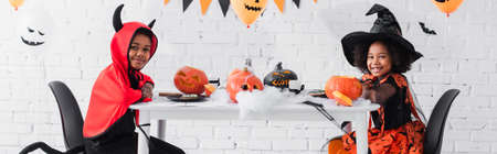 happy african american kids in halloween costumes sitting at table with pumpkins and decoration, bannerの写真素材