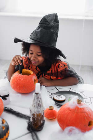 smiling african american girl in witch halloween costume carving pumpkinの写真素材