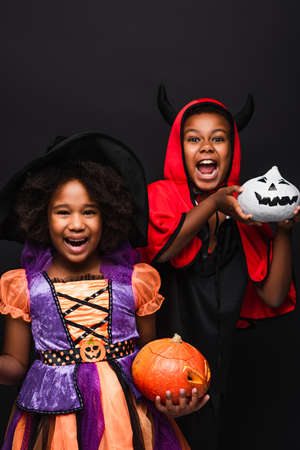cheerful african american children in halloween costumes holding spooky pumpkins isolated on blackの写真素材