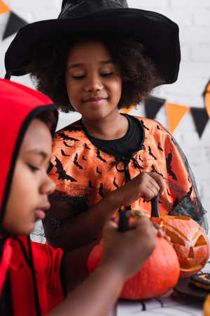 smiling african american girl in halloween costume looking at blurred brother drawing on pumpkinの写真素材
