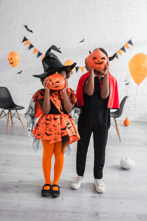 full length of african american kids in halloween costumes covering faces with carved pumpkins at homeの写真素材