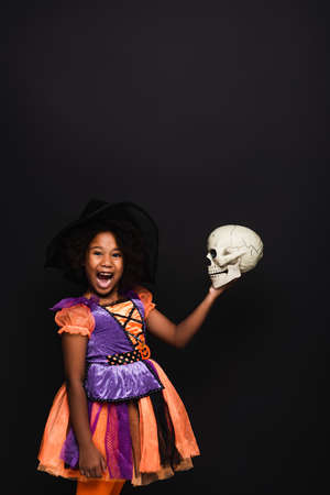 amazed african american girl in witch halloween costume holding skull isolated on blackの写真素材
