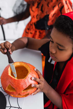 high angle view of african american boy in halloween costume carving pumpkin near blurred sisterの写真素材